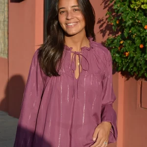 chica sonriendo delante de un muro de una casa con una blusa amplia rosa viejo con líneas de lúrex doradas y plateadas