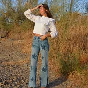 chica en el campo con un pantalón denim con parches de estrellas y blusa blanca manga larga con lúrex plateado